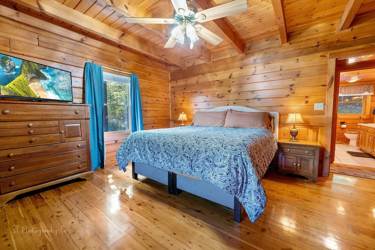 A comfortable bedroom with a large bed dressed in decorative bedding, a wooden dresser topped with a television, and soft blue curtains framing a window that lets in natural light. The overall vibe is calm and secluded, inviting relaxation and private downtime.