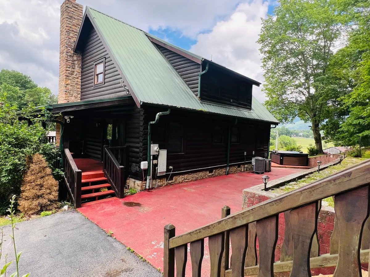 A welcoming exterior shot of the log cabin, featuring a green metal roof and a red driveway, nestled among lush trees. The inviting front porch hints at a perfect gathering spot before or after family adventures in the Smokies.