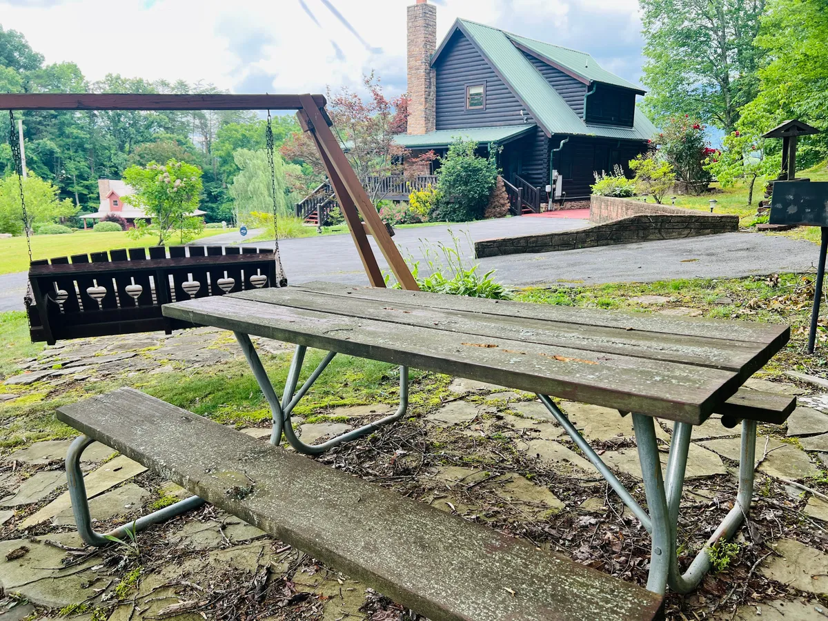 A picnic table and swing sit in a landscaped garden next to a classic log cabin house. The area is tranquil, with ample room for outdoor dining, playing, or simply enjoying the fresh air. The setting is ideal for picnics and gatherings with pets and family.