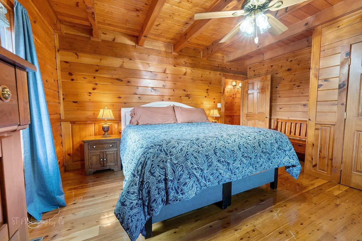 A bedroom with a patterned blue bedspread, wooden nightstands with lamps on each side, wooden walls and ceiling, and a ceiling fan. Sunlight filters in through the curtains, creating a tranquil and restful mood.
