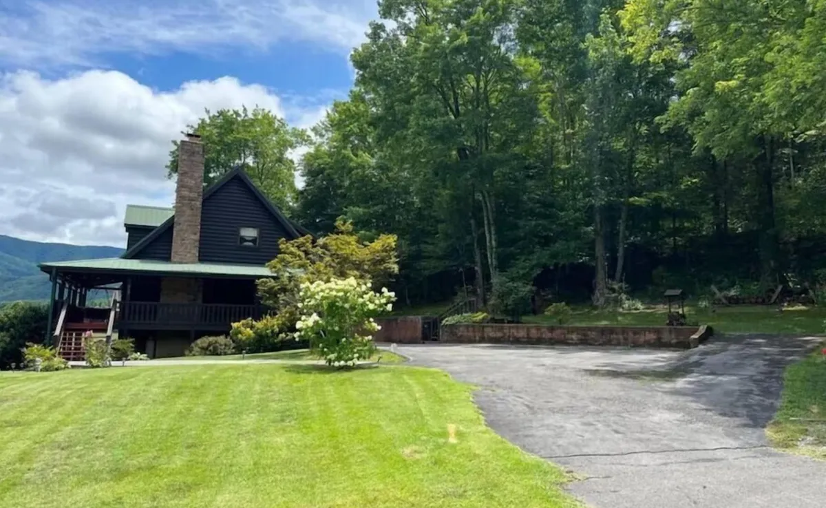 A broad exterior view of the property showcasing the welcoming front porch, landscaped yard, spacious driveway, and the cabin set against a backdrop of greenery. The porch invites relaxation, while the landscaped yard and driveway underscore practical amenities for guests arriving by car.