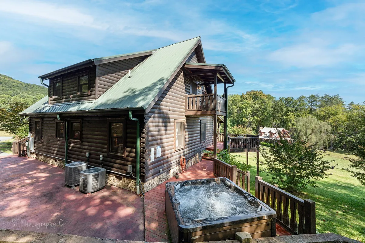 This exterior image showcases the log cabin's structure, a green roof, and a welcoming deck area with a hot tub. The deck overlooks the mountains, suggesting the scenic views and tranquil atmosphere described in the section. The hot tub is positioned to take in sunset and starlit views, perfectly capturing the inviting relaxation elements.