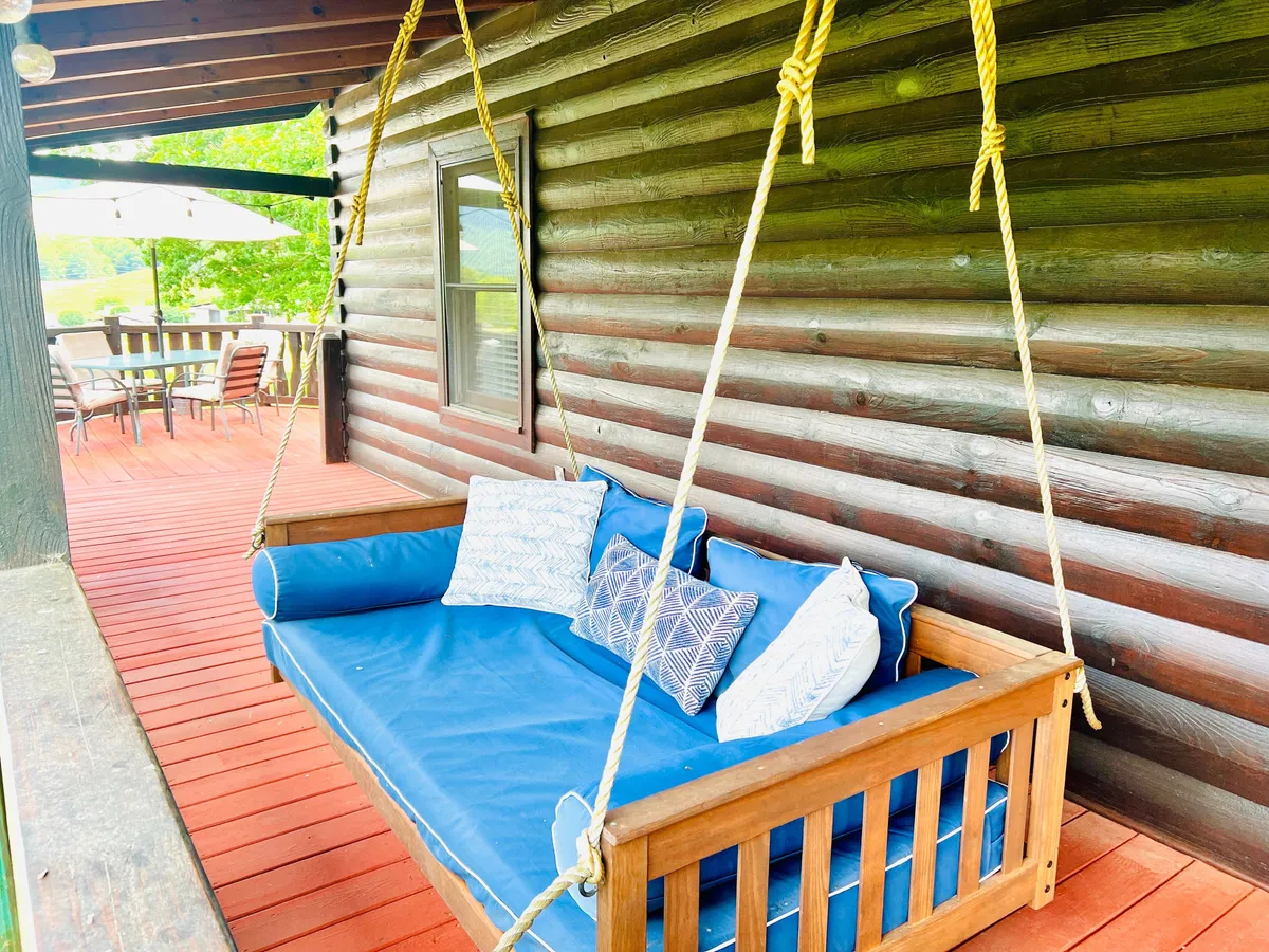 A serene exterior scene featuring a swinging couch on a wooden deck, an outdoor dining set, and beautiful sunset views. The space is designed for relaxation, reading, and enjoying meals outdoors, aligning with the section's mention of rocking chairs, porch swings, and al fresco dining.