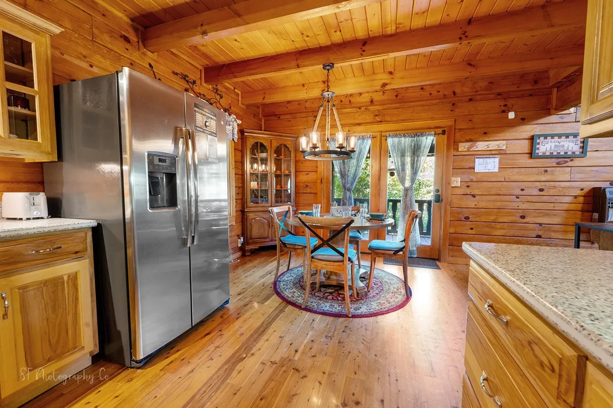 A wooden dining table for four sits beneath a chandelier, surrounded by large windows with curtains that draw in natural light. Stainless steel appliances are visible, tying the space to the kitchen, and the setting is ideal for intimate meals, board games, or morning coffee.