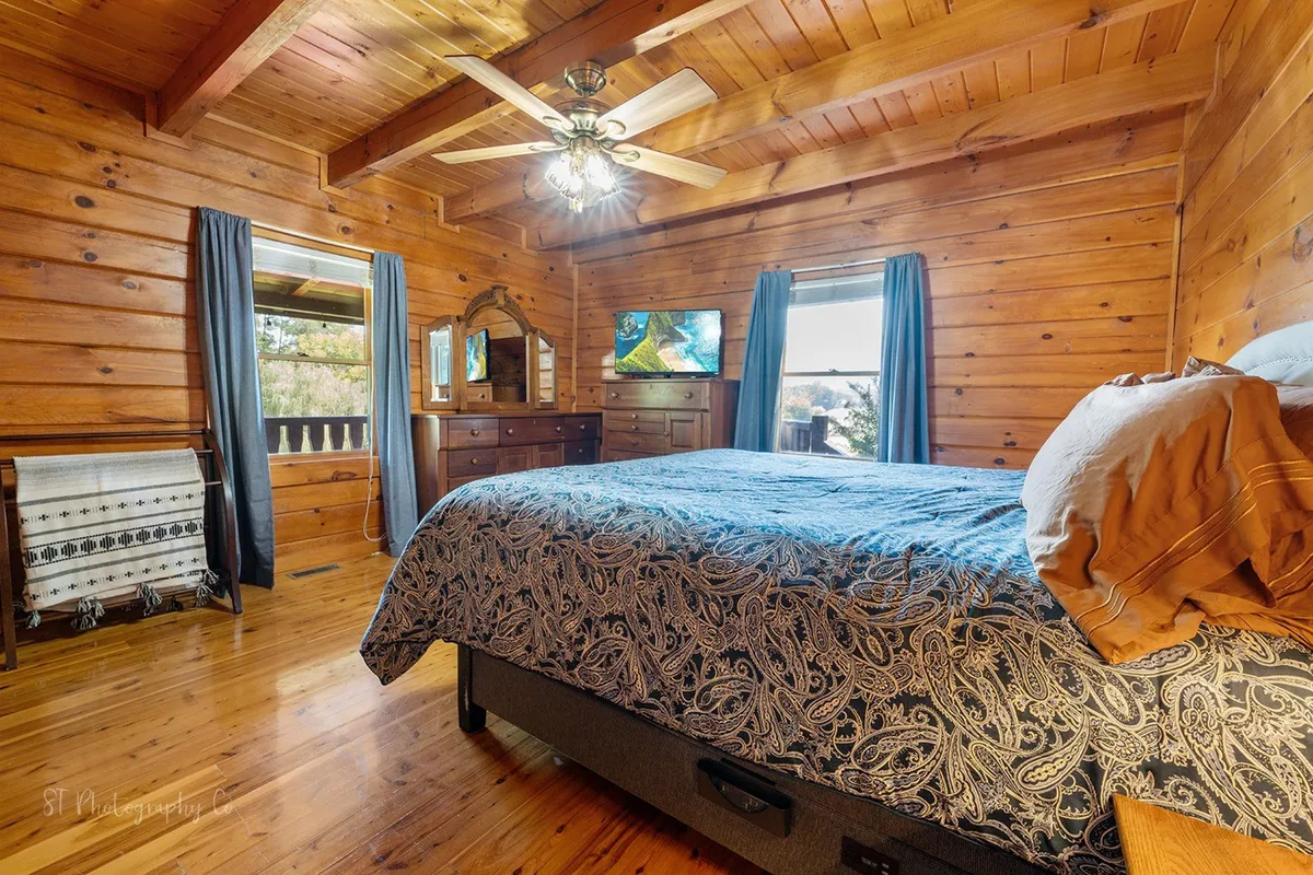A bedroom with wooden ceiling beams, a comfortable bed with patterned bedding, a large window offering a view of the outdoors, a wooden dresser with a mirror, and a ceiling fan. The atmosphere is serene and bright, with natural light streaming in.