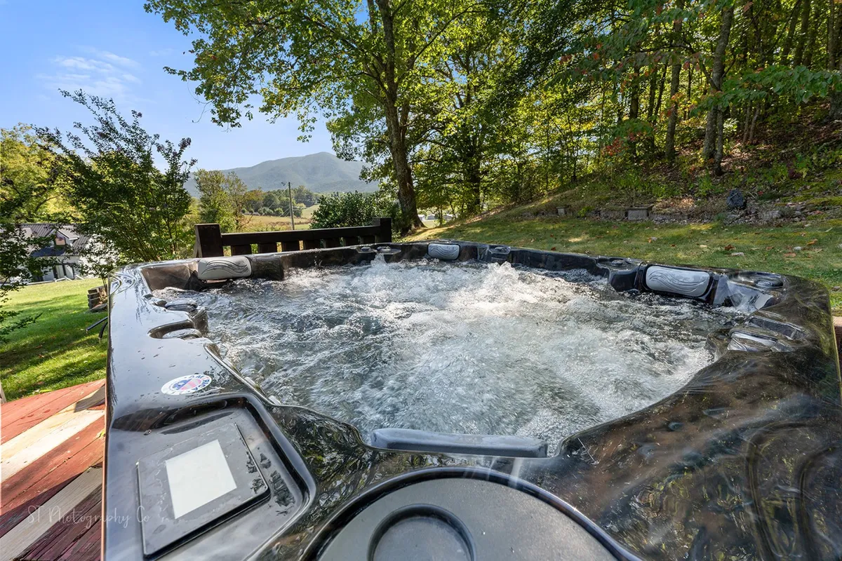 An inviting hot tub sits on a deck surrounded by greenery, with expansive mountain views in the background. The setting is tranquil, with ample space for several friends to relax together and enjoy nature.