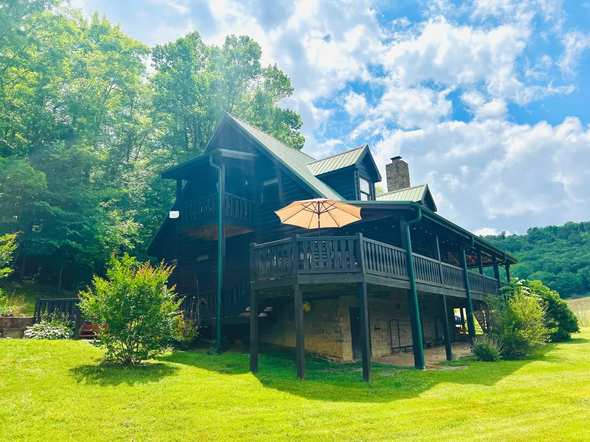 An exterior view of the cabin's wooden structure, green metal roof, and surrounding lush greenery, with an outdoor umbrella on the deck. The perspective situates the home within its natural setting and hints at both the proximity to nature and the welcoming, retreat-like atmosphere.