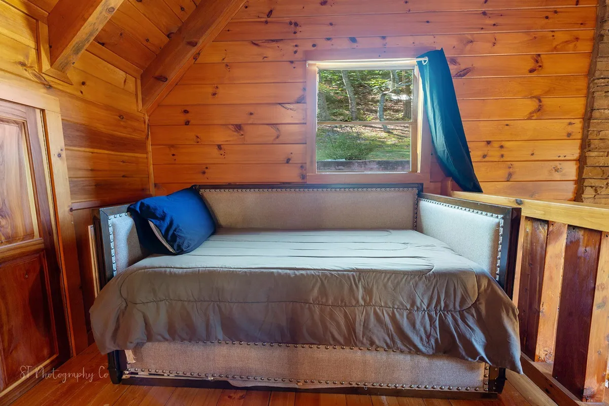 A cozy loft area with a daybed, large window, and wood-paneled walls. The daybed is styled for relaxation or sleepovers, and the window seat offers views and a perfect nook for daydreaming or reading.