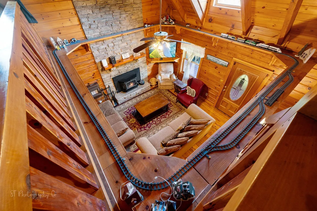 A unique view of the living space featuring a model train track circling above, the stone fireplace, comfortable seating area, and exposed wooden beams, creating a nostalgic and playful atmosphere ideal for family fun and celebrations.
